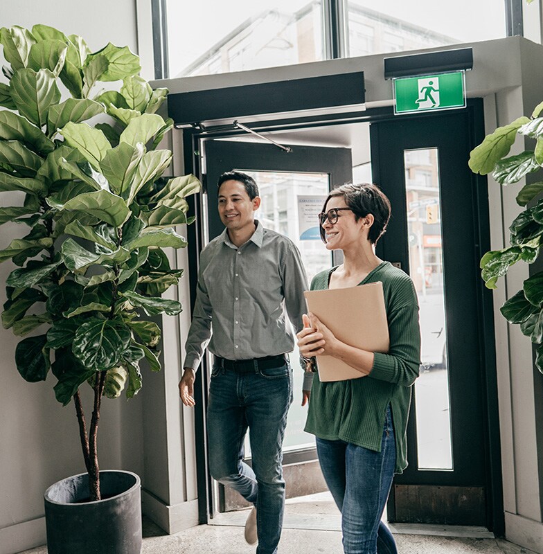 A man and a woman smiling and entering an office, with plants near the door
