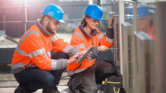 Two maintenance workers kneeling before a device, while one scrolls on a tablet